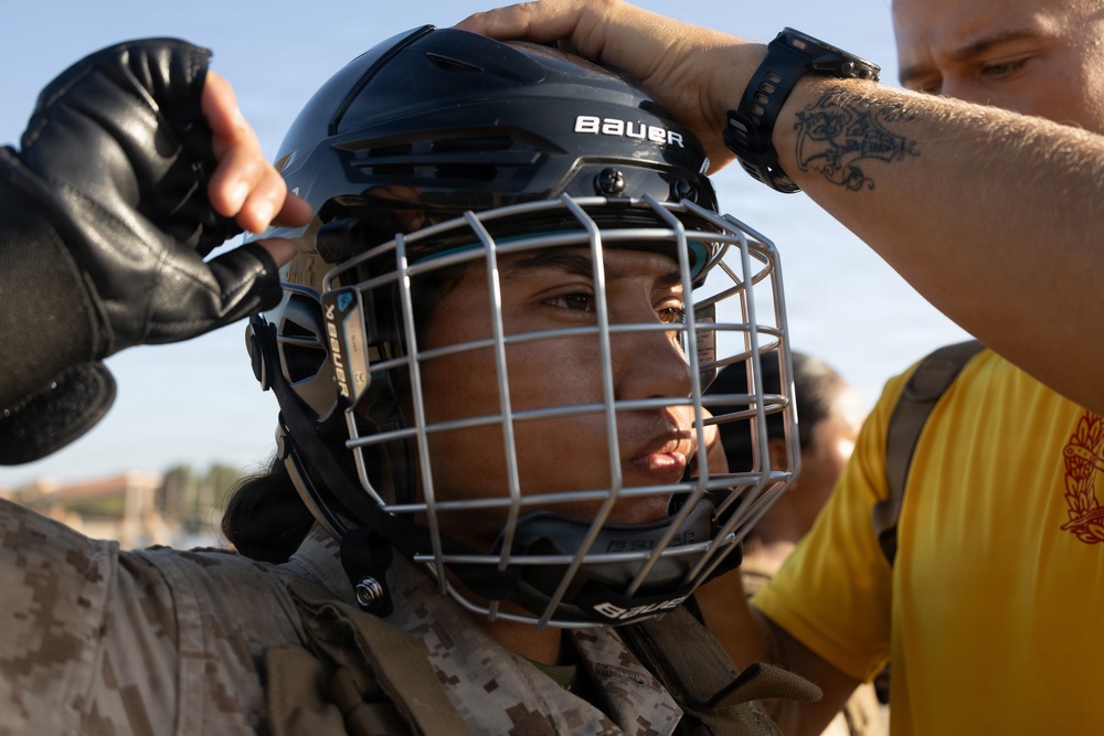 A person wearing U.S. Marine Corps desert camouflage uniform and black leather gloves is being fitted with a Bauer hockey-style protective helmet with a cage face guard. A second individual in a yellow shirt, wearing a watch and displaying a forearm tattoo, is adjusting the helmet from behind. The setting appears to be an outdoor training or combatives exercise environment.