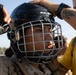 A person wearing U.S. Marine Corps desert camouflage uniform and black leather gloves is being fitted with a Bauer hockey-style protective helmet with a cage face guard. A second individual in a yellow shirt, wearing a watch and displaying a forearm tattoo, is adjusting the helmet from behind. The setting appears to be an outdoor training or combatives exercise environment.