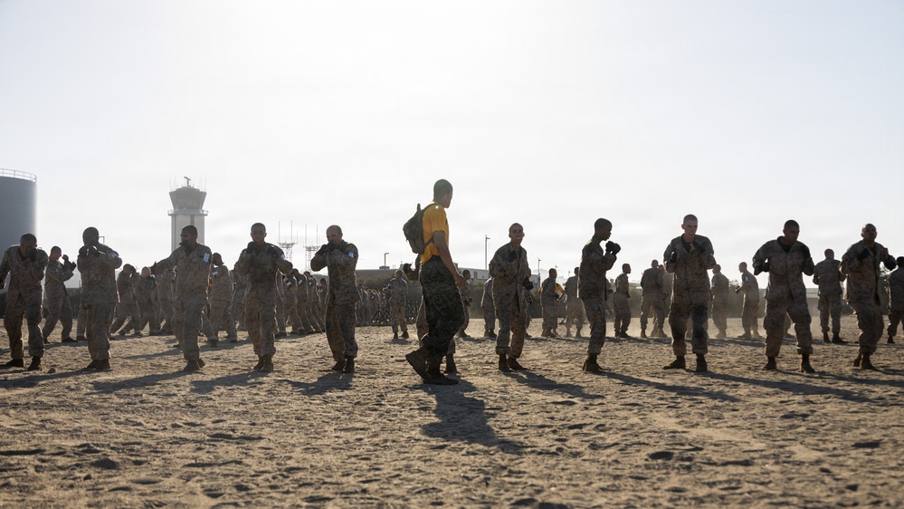 A large group of military personnel in camouflage uniforms are engaged in physical training or drill exercises on an open sandy/dirt field. One individual wearing a yellow shirt and carrying a backpack stands out prominently in the center foreground, likely serving as an instructor or observer. A control tower and industrial structures are visible in the background, suggesting a military airfield or base setting.