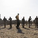 A large group of military personnel in camouflage uniforms are engaged in physical training or drill exercises on an open sandy/dirt field. One individual wearing a yellow shirt and carrying a backpack stands out prominently in the center foreground, likely serving as an instructor or observer. A control tower and industrial structures are visible in the background, suggesting a military airfield or base setting.