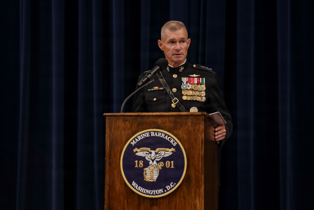 A senior U.S. Marine Corps officer in dress blues uniform stands at a wooden podium bearing the Marine Barracks Washington, D.C. seal (established 1801). The officer is delivering a speech or remarks at an official military ceremony or event. The formal setting, extensive medal rack, and aviation wings indicate a high-ranking officer at a significant official function.