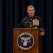 A senior U.S. Marine Corps officer in dress blues uniform stands at a wooden podium bearing the Marine Barracks Washington, D.C. seal (established 1801). The officer is delivering a speech or remarks at an official military ceremony or event. The formal setting, extensive medal rack, and aviation wings indicate a high-ranking officer at a significant official function.
