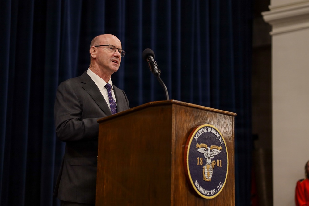 A man in a dark suit and purple tie is speaking at a wooden podium bearing the seal of Marine Barracks Washington, D.C., established in 1801. The setting appears to be a formal official event or ceremony held at the historic Marine Barracks facility. A blue curtain serves as the backdrop behind the speaker.
