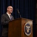 A man in a dark suit and purple tie is speaking at a wooden podium bearing the seal of Marine Barracks Washington, D.C., established in 1801. The setting appears to be a formal official event or ceremony held at the historic Marine Barracks facility. A blue curtain serves as the backdrop behind the speaker.