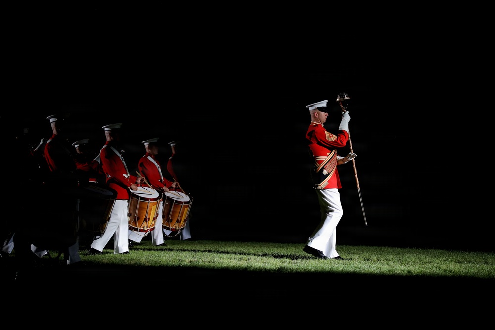 A military marching band performs a nighttime ceremony on a grass field, dramatically lit against a dark background. The lead figure, a drum major, marches ahead of the group holding a ceremonial mace and sword. The performers wear distinctive red tunics with white trousers and white peaked caps, consistent with U.S. Marine Corps dress uniform.