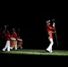 A military marching band performs a nighttime ceremony on a grass field, dramatically lit against a dark background. The lead figure, a drum major, marches ahead of the group holding a ceremonial mace and sword. The performers wear distinctive red tunics with white trousers and white peaked caps, consistent with U.S. Marine Corps dress uniform.