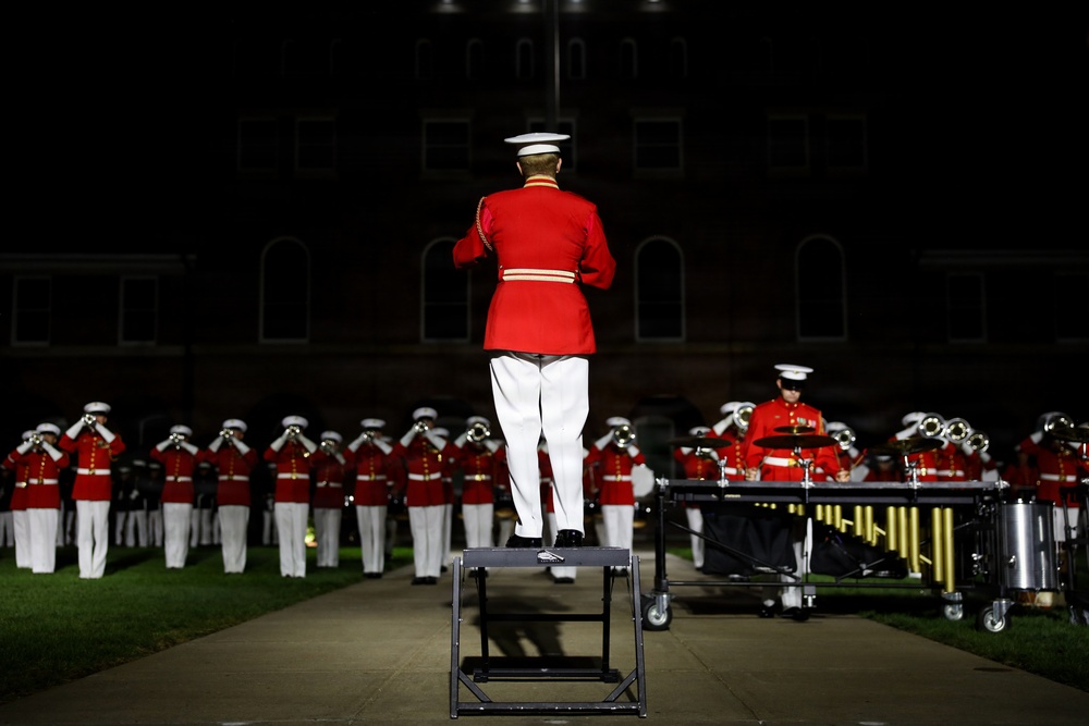 A military band conductor stands elevated on a podium, facing a large ensemble of musicians dressed in red and white dress uniforms performing at a nighttime outdoor ceremony. The band members are playing brass instruments and percussion in a formal parade ground setting. The dramatic lighting against the dark background highlights the precision and formality of the performance.