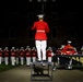 A military band conductor stands elevated on a podium, facing a large ensemble of musicians dressed in red and white dress uniforms performing at a nighttime outdoor ceremony. The band members are playing brass instruments and percussion in a formal parade ground setting. The dramatic lighting against the dark background highlights the precision and formality of the performance.