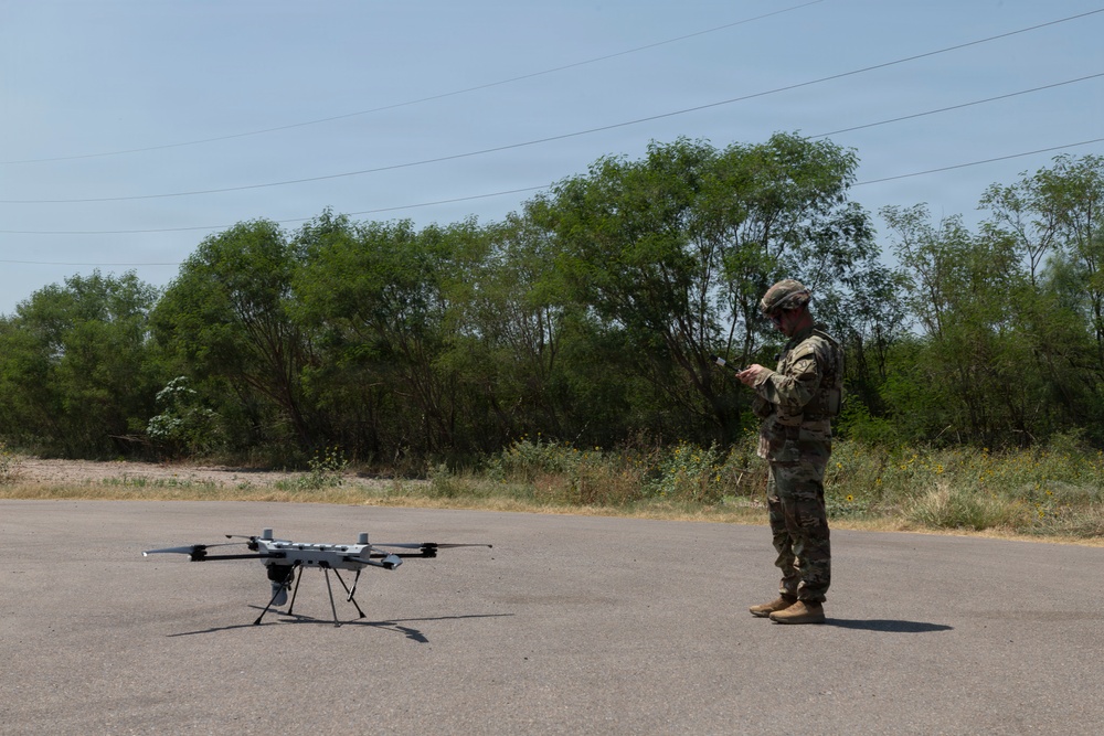 A military service member in camouflage uniform and helmet is operating a large multi-rotor drone on an open paved surface. The drone, which appears to be a heavy-lift or tactical unmanned aerial vehicle, is positioned on the ground while the operator uses a handheld controller. The setting appears to be an outdoor training or operational area with trees and brush in the background.