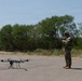 A military service member in camouflage uniform and helmet is operating a large multi-rotor drone on an open paved surface. The drone, which appears to be a heavy-lift or tactical unmanned aerial vehicle, is positioned on the ground while the operator uses a handheld controller. The setting appears to be an outdoor training or operational area with trees and brush in the background.