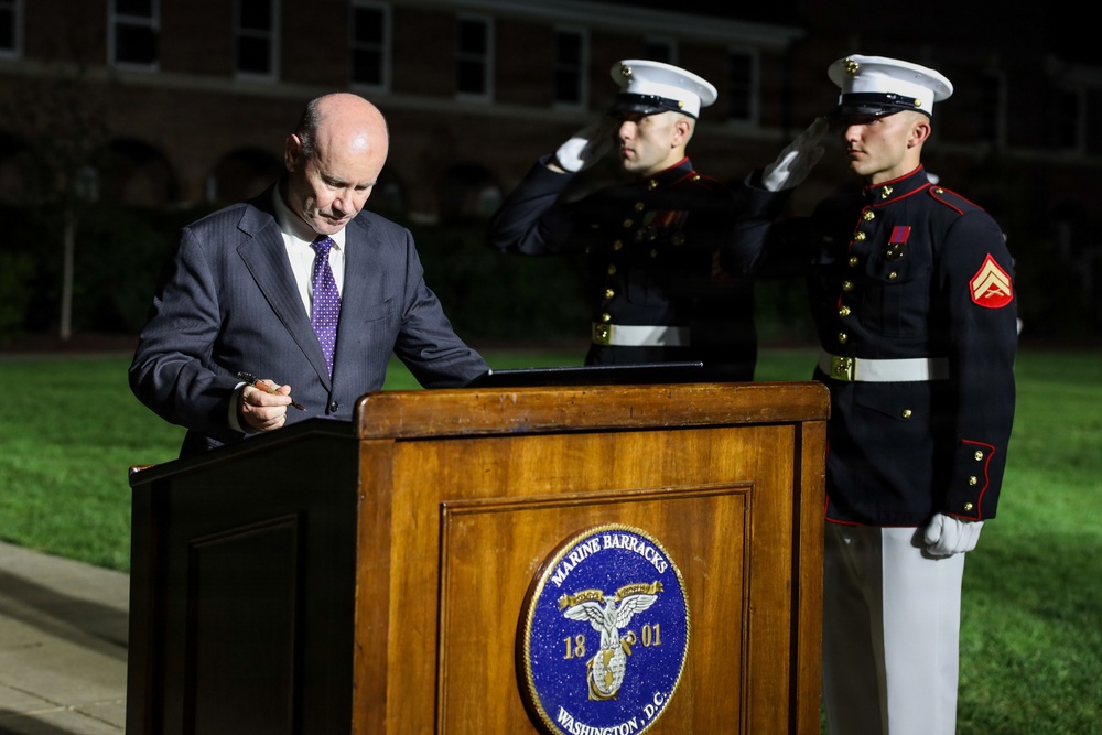 A civilian in a dark suit stands at a wooden podium bearing the Marine Barracks Washington D.C. seal (established 1801), appearing to sign or review a document. Two U.S. Marines in dress blue uniforms stand at attention behind him, one executing a salute. The event appears to be a formal military ceremony taking place at night on the Marine Barracks Washington parade ground.