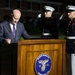 A civilian in a dark suit stands at a wooden podium bearing the Marine Barracks Washington D.C. seal (established 1801), appearing to sign or review a document. Two U.S. Marines in dress blue uniforms stand at attention behind him, one executing a salute. The event appears to be a formal military ceremony taking place at night on the Marine Barracks Washington parade ground.