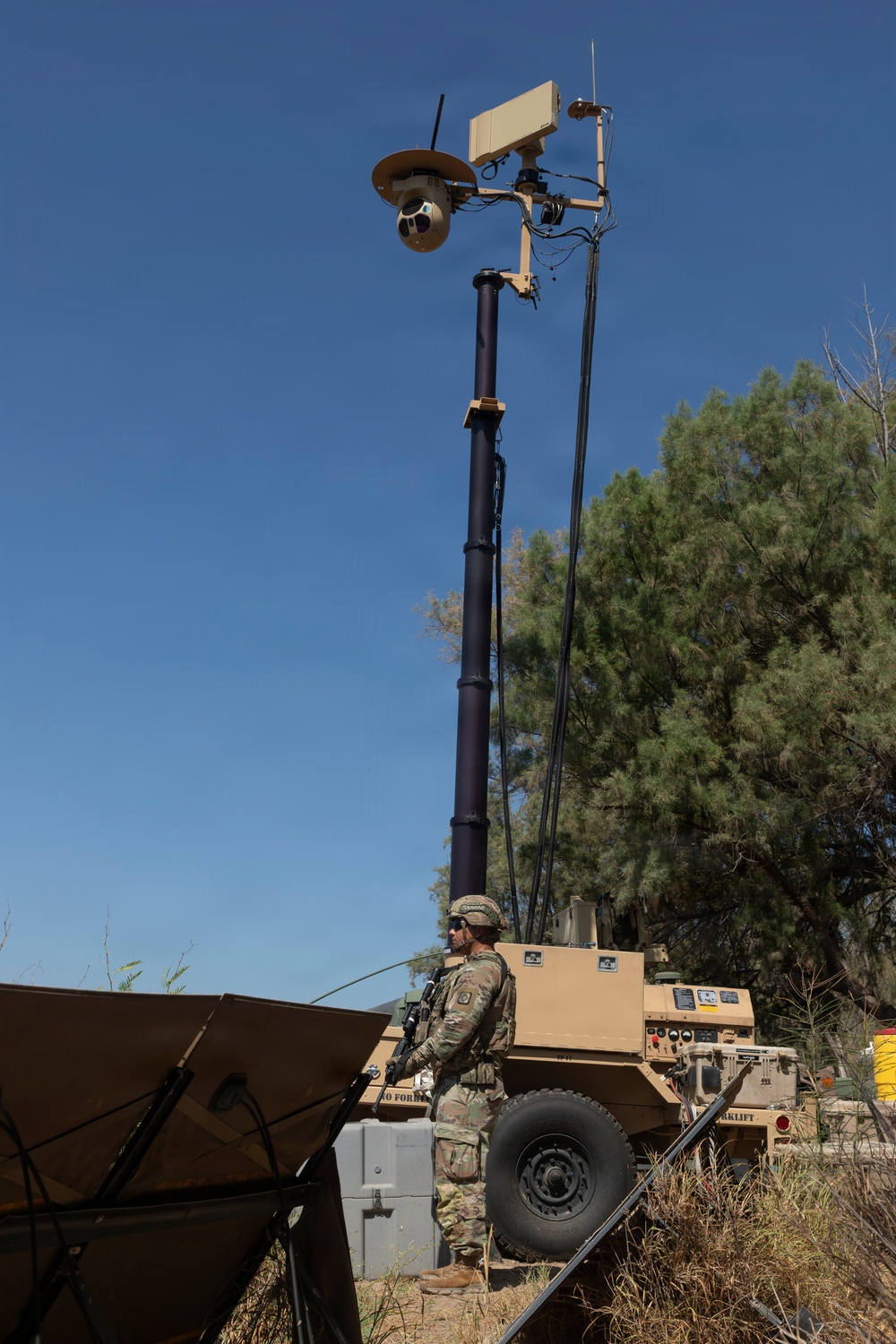 A U.S. Army soldier in full combat gear stands beside a military surveillance tower mounted on a wheeled trailer unit. The tower features a pan-tilt-zoom (PTZ) camera system and communications equipment elevated on an extendable mast. The scene appears to be a field exercise or forward operating base setup in an arid, semi-wooded environment.