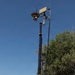 A U.S. Army soldier in full combat gear stands beside a military surveillance tower mounted on a wheeled trailer unit. The tower features a pan-tilt-zoom (PTZ) camera system and communications equipment elevated on an extendable mast. The scene appears to be a field exercise or forward operating base setup in an arid, semi-wooded environment.