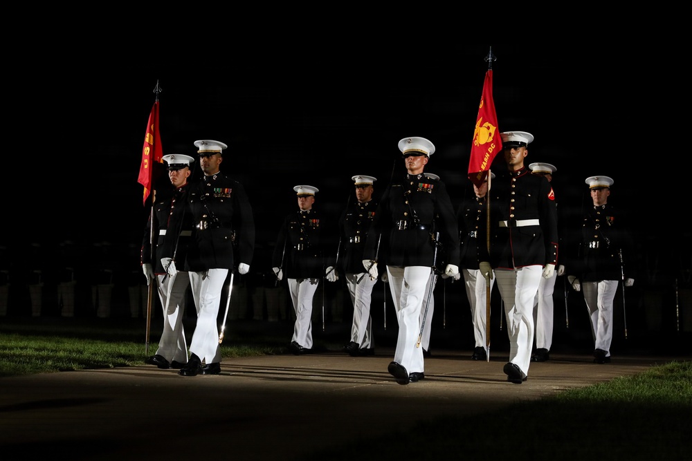 A group of United States Marines in dress blue uniforms are marching in formation during a nighttime ceremonial parade. They carry red Marine Corps guidon flags and wear white trousers, white gloves, and white peaked caps. The scene is illuminated against a dark background, consistent with an evening parade or sunset ceremony.