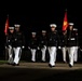 A group of United States Marines in dress blue uniforms are marching in formation during a nighttime ceremonial parade. They carry red Marine Corps guidon flags and wear white trousers, white gloves, and white peaked caps. The scene is illuminated against a dark background, consistent with an evening parade or sunset ceremony.