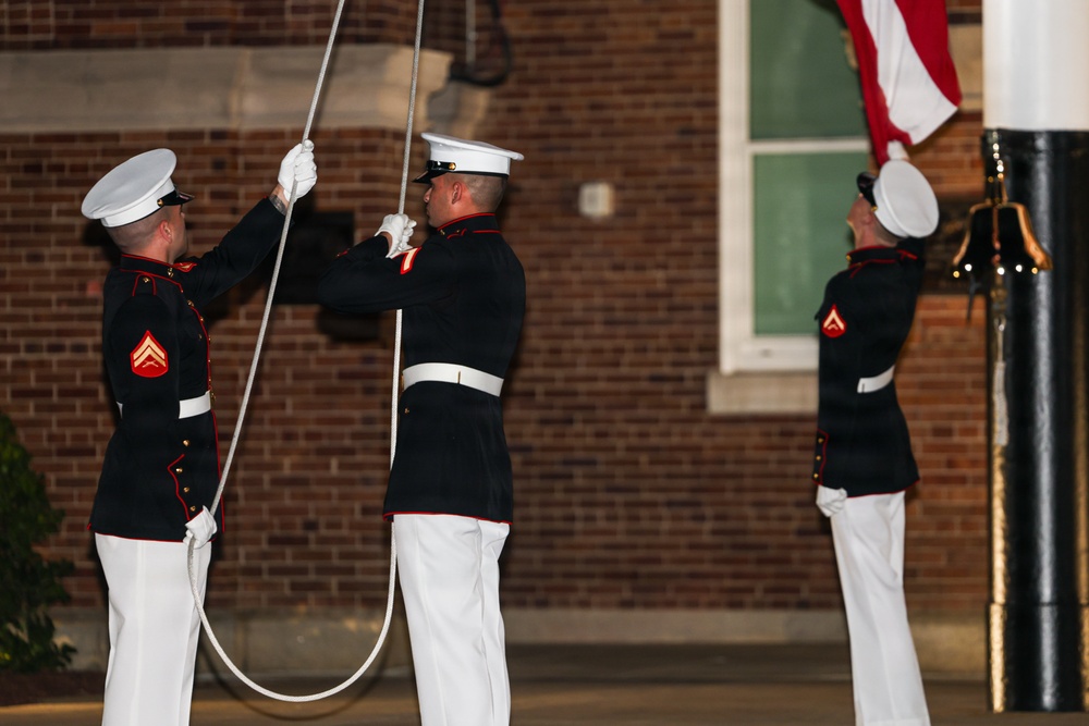 Three U.S. Marines in dress blues are performing a formal flag ceremony, likely a retreat or evening colors ceremony. They are wearing their distinctive dress uniform consisting of dark blue coats with red trim and white trousers, white covers, and white gloves. Two Marines are handling the halyard rope while a third manages the flag at the base of a flagpole.