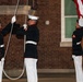 Three U.S. Marines in dress blues are performing a formal flag ceremony, likely a retreat or evening colors ceremony. They are wearing their distinctive dress uniform consisting of dark blue coats with red trim and white trousers, white covers, and white gloves. Two Marines are handling the halyard rope while a third manages the flag at the base of a flagpole.