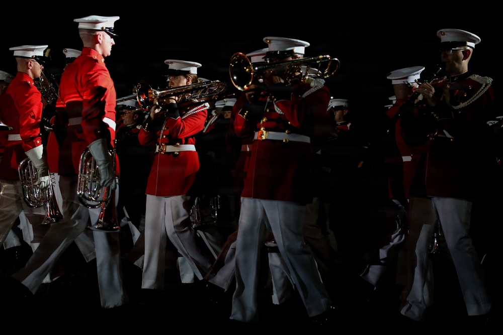 A military marching band is performing in formation under dramatic low-key lighting against a dark background. The musicians are dressed in distinctive red tunics with white trousers and white peaked caps, consistent with U.S. Marine Corps dress uniform. They are actively playing brass instruments while marching in coordinated formation.