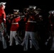 A military marching band is performing in formation under dramatic low-key lighting against a dark background. The musicians are dressed in distinctive red tunics with white trousers and white peaked caps, consistent with U.S. Marine Corps dress uniform. They are actively playing brass instruments while marching in coordinated formation.