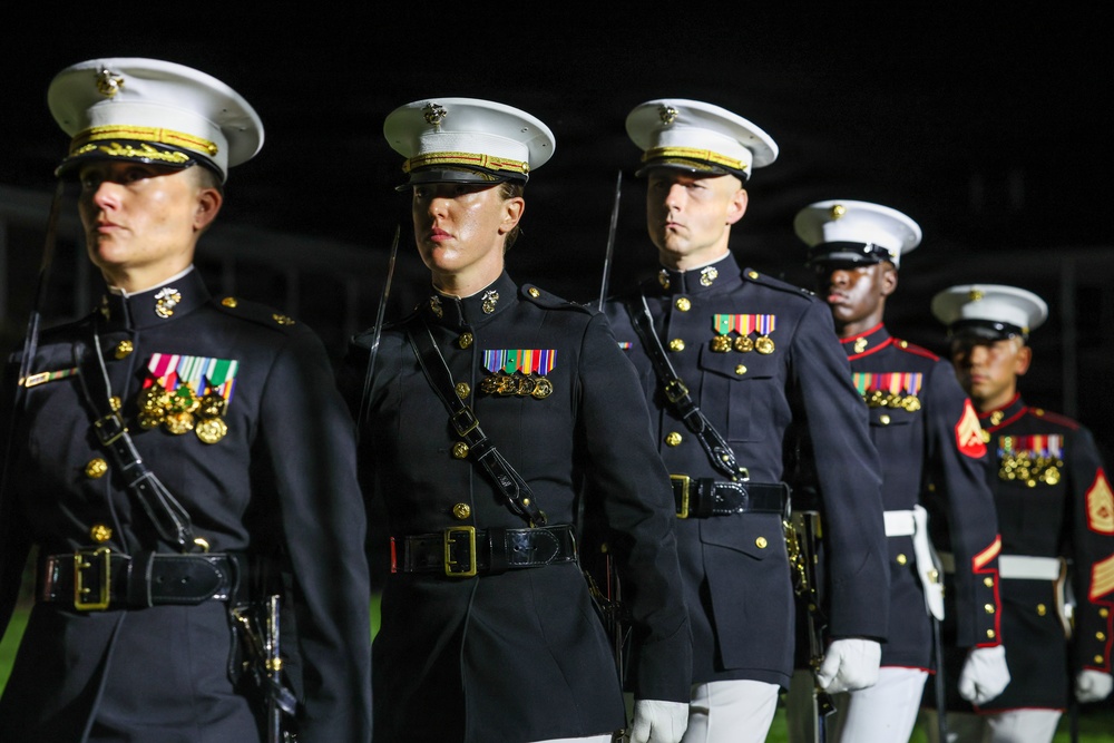 A group of United States Marine Corps personnel are standing in formal dress blue uniforms during what appears to be a ceremonial parade or evening parade event. The Marines are in precise formation, wearing their distinctive dress blues with white covers (caps), medals, and ceremonial equipment. The dark background suggests this is a nighttime military ceremony.