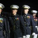 A group of United States Marine Corps personnel are standing in formal dress blue uniforms during what appears to be a ceremonial parade or evening parade event. The Marines are in precise formation, wearing their distinctive dress blues with white covers (caps), medals, and ceremonial equipment. The dark background suggests this is a nighttime military ceremony.