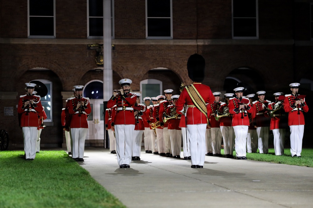 A military marching band performs at night in front of a brick building, likely during a formal ceremonial event. The musicians are dressed in distinctive red tunics with white trousers and white peaked caps, consistent with U.S. Marine Corps Band dress uniforms. A drum major or conductor figure wearing a tall black bearskin hat leads the formation.