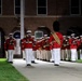 A military marching band performs at night in front of a brick building, likely during a formal ceremonial event. The musicians are dressed in distinctive red tunics with white trousers and white peaked caps, consistent with U.S. Marine Corps Band dress uniforms. A drum major or conductor figure wearing a tall black bearskin hat leads the formation.