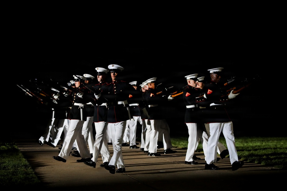 A precision drill team, identifiable as U.S. Marines based on their distinctive dress uniforms, performs a nighttime ceremonial drill routine. The Marines are marching in tight formation while executing synchronized rifle movements. The dramatic lighting against a dark background highlights the precision and discipline of the performance.