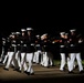 A precision drill team, identifiable as U.S. Marines based on their distinctive dress uniforms, performs a nighttime ceremonial drill routine. The Marines are marching in tight formation while executing synchronized rifle movements. The dramatic lighting against a dark background highlights the precision and discipline of the performance.