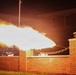 A nighttime military ceremonial cannon firing is captured, producing a large muzzle blast and flame extending horizontally from behind a brick wall. Two uniformed personnel, appearing to be U.S. Marines based on their distinctive white dress covers, are visible standing at attention on the right side of the frame. The dramatic long-exposure or high-speed photograph captures the full extent of the propellant flame against the dark night sky.