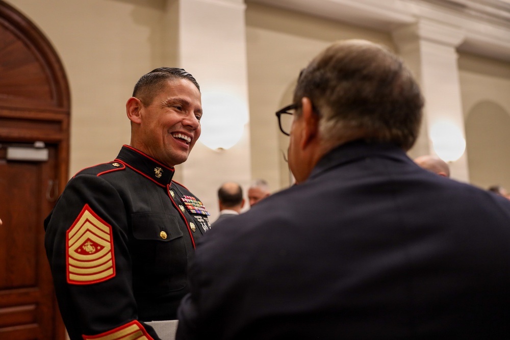 A U.S. Marine Corps Sergeant Major in dress blues uniform is engaged in a cheerful conversation with a civilian in a formal indoor setting. The Marine is smiling broadly and wearing multiple service ribbons and medals. The setting appears to be a formal reception or ceremony in an ornate building.