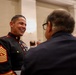 A U.S. Marine Corps Sergeant Major in dress blues uniform is engaged in a cheerful conversation with a civilian in a formal indoor setting. The Marine is smiling broadly and wearing multiple service ribbons and medals. The setting appears to be a formal reception or ceremony in an ornate building.