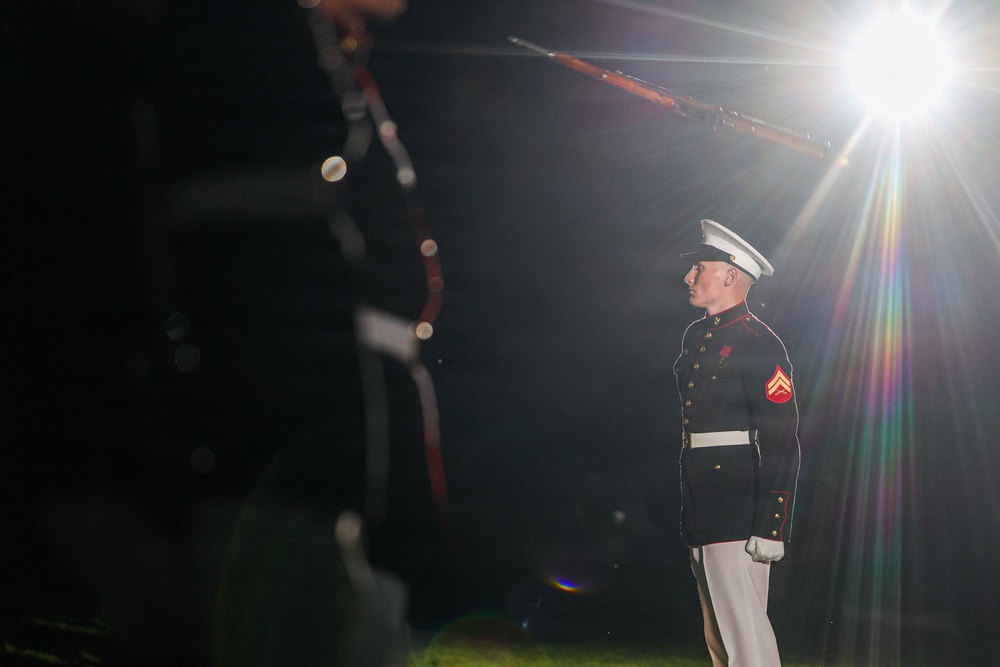 A U.S. Marine in dress blues uniform stands at attention during what appears to be a formal ceremonial event at night. The Marine is illuminated by a bright spotlight creating a dramatic lens flare effect. In the foreground and background, rifle drill movements are partially visible, suggesting a Silent Drill Platoon or similar precision drill performance.