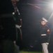 A U.S. Marine in dress blues uniform stands at attention during what appears to be a formal ceremonial event at night. The Marine is illuminated by a bright spotlight creating a dramatic lens flare effect. In the foreground and background, rifle drill movements are partially visible, suggesting a Silent Drill Platoon or similar precision drill performance.