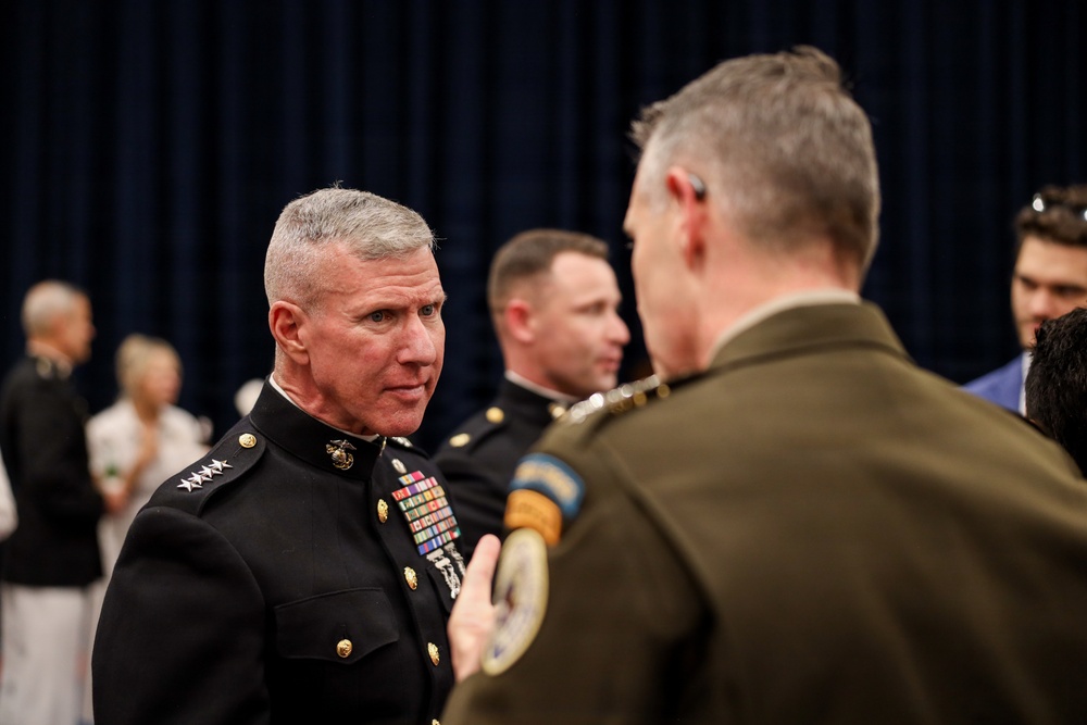 A formal military gathering featuring uniformed personnel in conversation. The central figure wears a U.S. Marine Corps dress blue uniform with four-star rank insignia and multiple service ribbons, indicating a General officer rank. The setting appears to be an official ceremony or reception with a dark curtained backdrop.