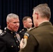 A formal military gathering featuring uniformed personnel in conversation. The central figure wears a U.S. Marine Corps dress blue uniform with four-star rank insignia and multiple service ribbons, indicating a General officer rank. The setting appears to be an official ceremony or reception with a dark curtained backdrop.