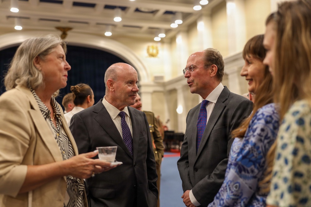 A formal reception or networking event taking place in an elegant, well-lit hall with classical architectural features. Four individuals are engaged in conversation in the foreground, with two men in dark suits and two women, while additional attendees are visible in the background. The setting suggests a diplomatic, governmental, or institutional gathering.