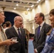 A formal reception or networking event taking place in an elegant, well-lit hall with classical architectural features. Four individuals are engaged in conversation in the foreground, with two men in dark suits and two women, while additional attendees are visible in the background. The setting suggests a diplomatic, governmental, or institutional gathering.