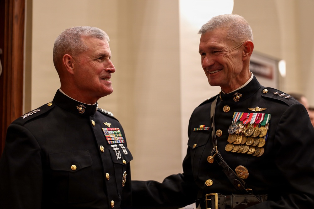 Two senior United States Marine Corps officers in dress blue uniforms are engaged in a friendly conversation, smiling at each other. Both display extensive medal ribbons and rank insignia consistent with general officer grades. The setting appears to be a formal military ceremony or official event.