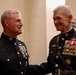 Two senior United States Marine Corps officers in dress blue uniforms are engaged in a friendly conversation, smiling at each other. Both display extensive medal ribbons and rank insignia consistent with general officer grades. The setting appears to be a formal military ceremony or official event.