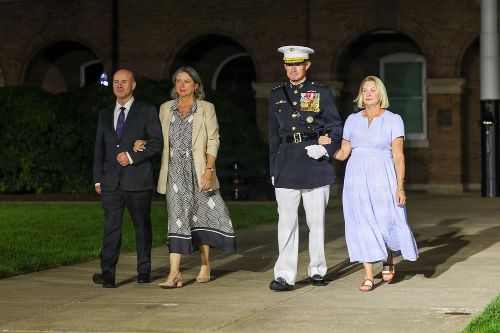 Four adults are walking together on a paved pathway at night in front of a brick building with arched architecture. One individual is wearing a U.S. Marine Corps dress uniform with medals, white gloves, and a white-topped service cap. The group appears to be attending or departing a formal military ceremony or event.