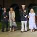 Four adults are walking together on a paved pathway at night in front of a brick building with arched architecture. One individual is wearing a U.S. Marine Corps dress uniform with medals, white gloves, and a white-topped service cap. The group appears to be attending or departing a formal military ceremony or event.