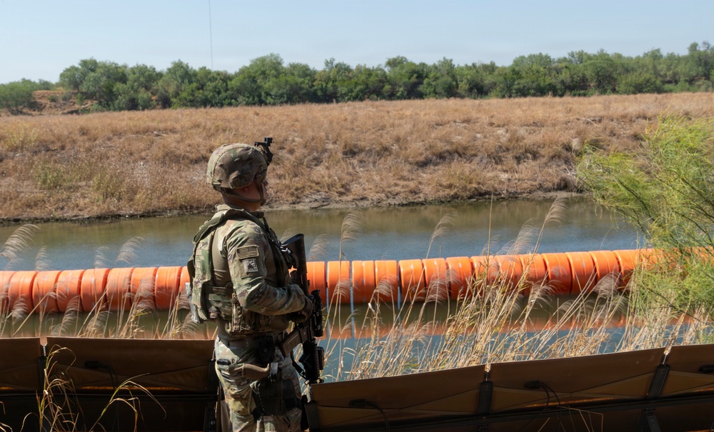 A uniformed soldier in full combat gear stands watch over a river barrier consisting of large orange buoys or floating barriers stretching across a waterway. The scene appears to be a border security operation, with dry grassland and dense tree line visible on the opposite bank. The soldier is armed and equipped with tactical gear including a helmet with mounted device.