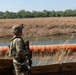 A uniformed soldier in full combat gear stands watch over a river barrier consisting of large orange buoys or floating barriers stretching across a waterway. The scene appears to be a border security operation, with dry grassland and dense tree line visible on the opposite bank. The soldier is armed and equipped with tactical gear including a helmet with mounted device.
