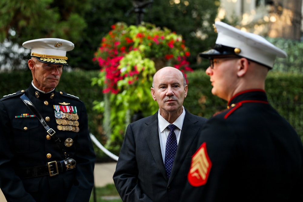 Three individuals are gathered outdoors in what appears to be a formal military or ceremonial setting. Two are wearing United States Marine Corps dress uniforms, while one civilian in a dark suit and tie stands between them. The setting includes decorative floral arrangements and manicured hedges in the background.