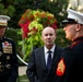 Three individuals are gathered outdoors in what appears to be a formal military or ceremonial setting. Two are wearing United States Marine Corps dress uniforms, while one civilian in a dark suit and tie stands between them. The setting includes decorative floral arrangements and manicured hedges in the background.