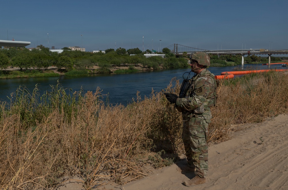 A soldier in full camouflage military uniform stands on a sandy riverbank, holding a device and monitoring the waterway. A line of orange floating barriers is visible stretching across the river in the background. A bridge and urban structures are visible on the opposite bank.