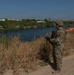 A soldier in full camouflage military uniform stands on a sandy riverbank, holding a device and monitoring the waterway. A line of orange floating barriers is visible stretching across the river in the background. A bridge and urban structures are visible on the opposite bank.
