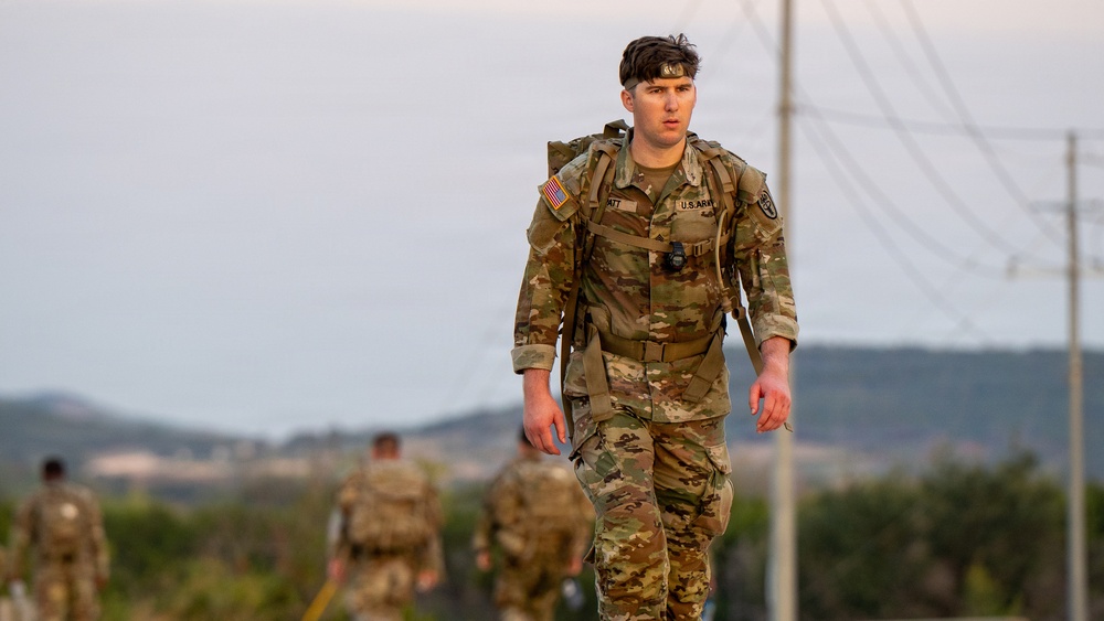 A U.S. Army soldier in Operational Camouflage Pattern (OCP) uniform is conducting a ruck march, carrying a loaded rucksack. The soldier is in the foreground and is clearly identifiable by 'U.S. ARMY' chest tape and an American flag patch on the left shoulder. Additional soldiers in similar uniforms are visible in the blurred background, suggesting a group training exercise.